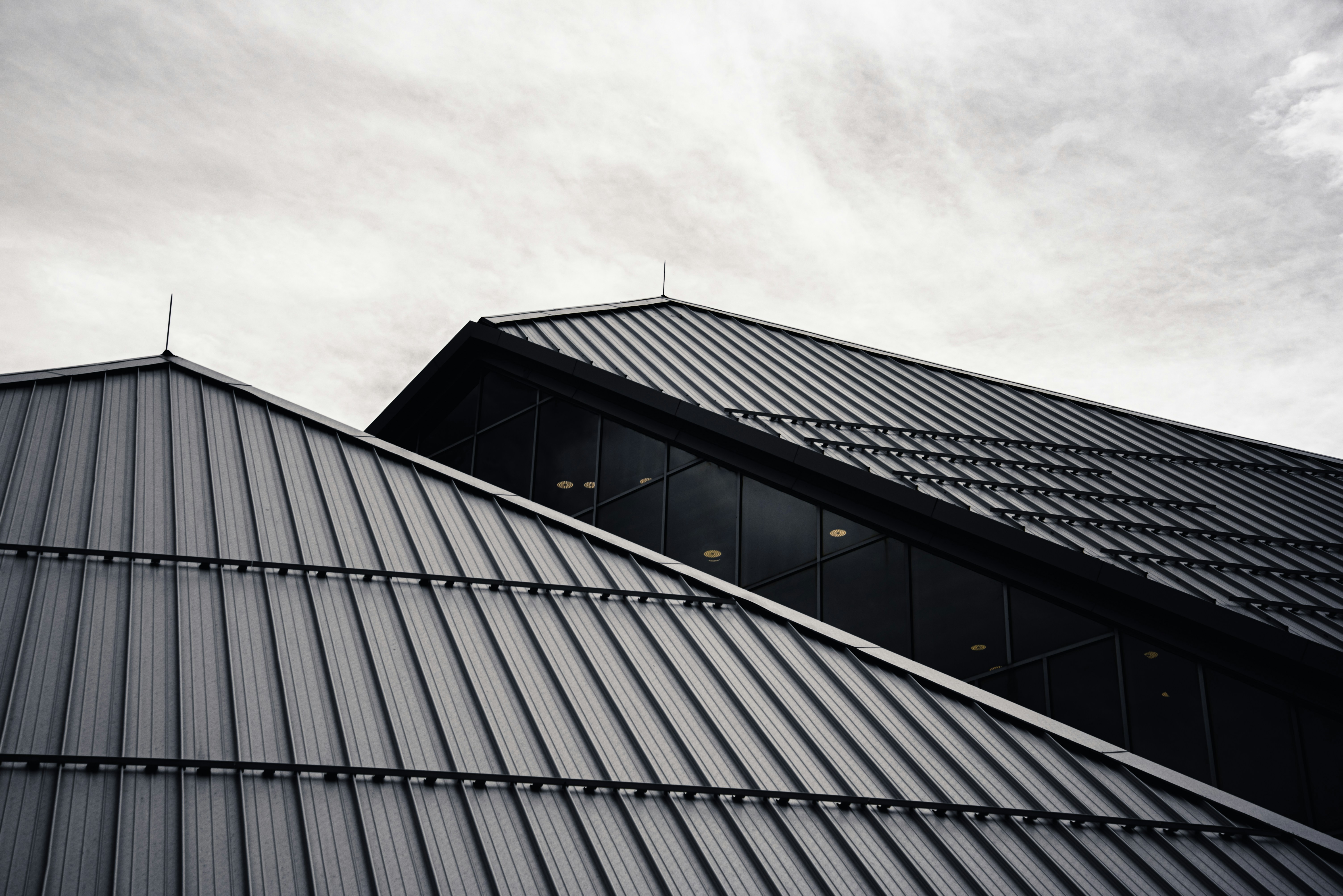 Upward view of a black standing-seam metal roof with a glass clerestory window along the ridge, set against an overcast sky