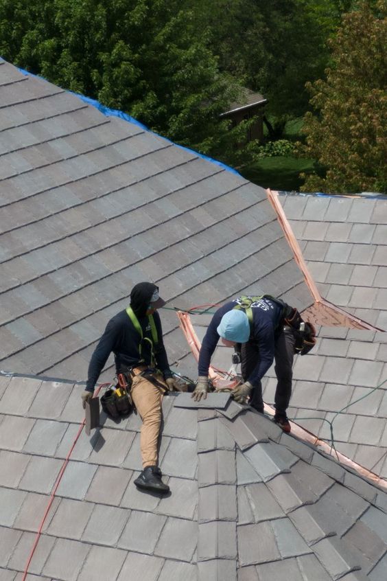 Two roofers in safety harnesses installing roof tiles at a ridge junction, working at height