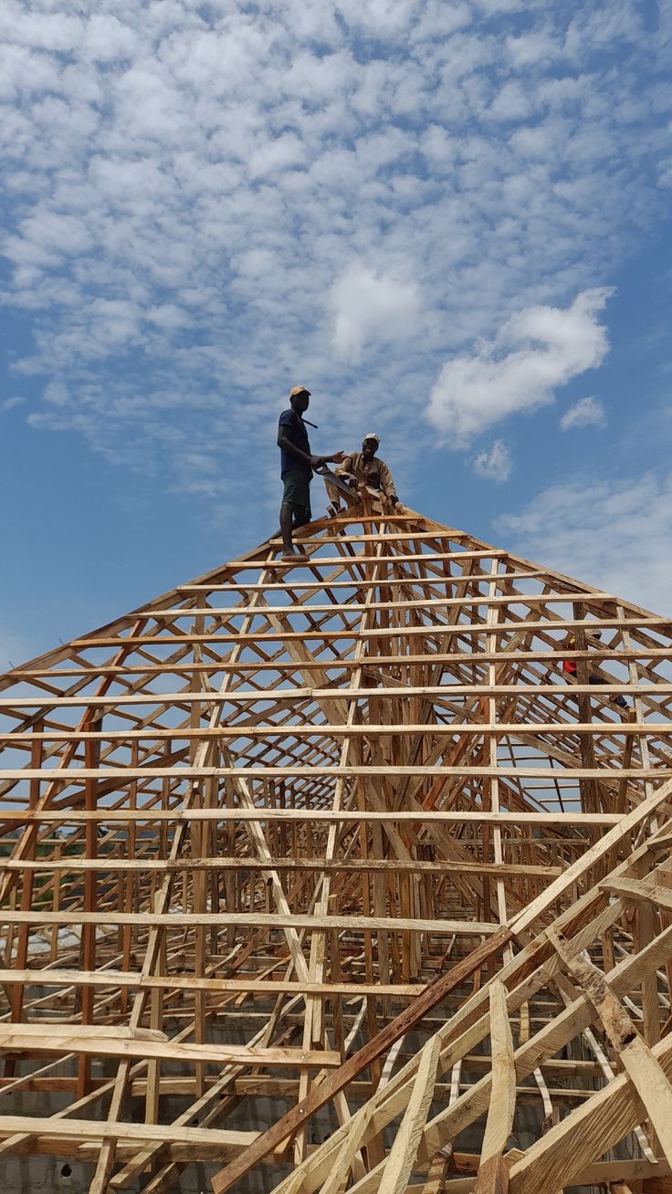 Two roofers standing atop a large timber roof truss frame during construction, working at height against a clear sky