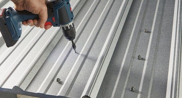 Installer using a power drill to fasten a self-drilling screw into a corrugated aluminium roofing sheet